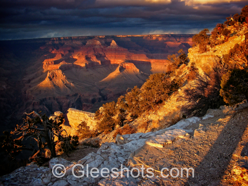 A vibrant sunset illuminates the Grand Canyon, highlighting its layered rock formations and rugged terrain. Scattered clouds hover above while sunlight enhances the deep reds and oranges of the canyon walls.  Arizona Fine Art Landscape Photography Print for Sale.