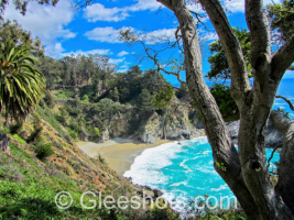 Scenic coastal view of McWay Falls cascading onto a sandy beach in Julia Pfeiffer Burns State Park in California. The turquoise ocean meets the shore, framed by lush green trees and vegetation under a bright blue sky with white clouds along the coast of Big Sur.  California Fine Art Landscape Photography Print for Sale.