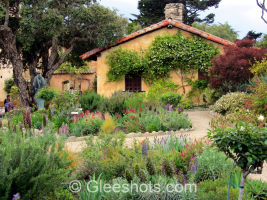 In this view of the Carmel Mission in California, a charming cottage with a red-tiled roof is surrounded by a lush garden full of diverse plants and colorful flowers. A gravel pathway winds through the vibrant greenery. Creeping vines adorn the cottage walls,  and a statue can be seen in amongst the trees.  California Fine Art Landscape Photography Print for Sale.