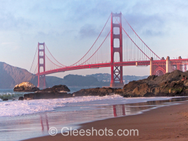 Image of the Golden Gate Bridge in San Francisco glowing red at sunset, with rocky shores in the foreground. The bridge is bathed in soft sunlight and its reflection can be seen in the beach below, with patches of fog in a partially clear sky above.  California Fine Art Landscape Photography Print for Sale.
