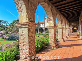 A view of the historic San Juan Capistrano mission with a red-tiled walkway and rustic brick arches, overlooking a vibrant garden with lush greenery and blooming flowers. In the background, Spanish-style buildings and a bell tower are visible under a clear blue sky.  California Fine Art Landscape Photography Print for Sale.