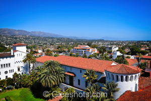 A scenic view of Santa Barbara from the courthouse tower, featuring a mix of white red-roofed buildings, palm trees, and green spaces. The backdrop includes a mountain range under a clear blue sky with the ocean in the distance.   California Fine Art Landscape Photography Print for Sale.