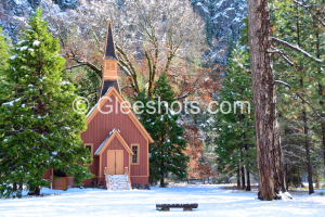 The Yosemite Valley Chapel is surrounded by snow-covered trees in a winter landscape. Snow blankets the ground, and the trees display a mix of green foliage and bare branches. A wooden bench can be seen in the foreground.   National Park Fine Art Landscape Photography Print for Sale.