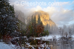 El Capitan in Yosemite is featured in this majestic winter sunset scene.  The golden mountain peaks of El Capitan are bathed by sunlight and are partially shrouded in clouds and fog.  A snow-covered forest is shown in the foreground, with the Merced river running through the scene.   National Park Fine Art Landscape Photography Print for Sale.