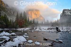 El Capitan is featured in this serene winter landscape sunset scene, with the Merced river and snow-covered rocks in the foreground.  Snow-dusted trees can be seen as mist rises in the distance against a backdrop of majestic mountains bathed in golden sunlight.   Yosemite National Park Fine Art Landscape Photography Print for Sale.