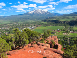 A scenic view of Carbondale, Colorado, taken from a red-orange rocky ledge. A snow-capped Mount Sopris rises prominently in the background under a bright blue sky with scattered clouds.   Colorado Fine Art Landscape Photography Print for Sale.
