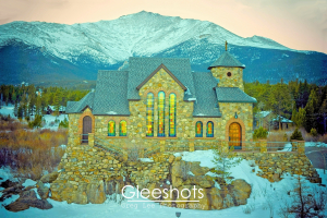 The stone Chapel on the Rock in Allenspark, Colorado, with colorful stained glass windows, sits on a rocky hill, surrounded by a snowy landscape and pine trees. In the background, the snow-capped Rocky mountains are visible under a softly lit evening sky.  Colorado Fine Art Landscape Photography Print for Sale.