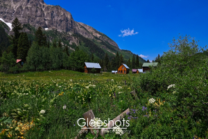 In Gothic Colorado, a lush green meadow with wildflowers, a wooden fence,  and cabins sit beneath Gothic Mountain under a bright blue sky.  Colorado Fine Art Landscape Photography for Sale.