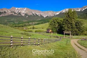 Along Last Dollar Road near Ridgway, Colorado,  this scenic landscape features a winding dirt road leading to a wooden cabin surrounded by lush green fields and tall trees. A wooden fence runs parallel to the road, and the snow-capped San Juan mountains rise in the background under a clear blue sky.  Colorado Landscape Photography Print for Sale.