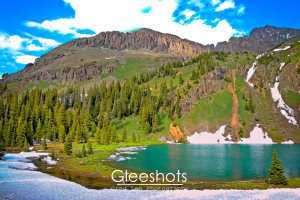Lower Blue Lake is an alpine lake with turquoise water, shown here surrounded by pine trees, patches of snow, and green hills. Rugged mountain peaks rise in the background.  Colorado Fine Art Landscape Photography for Sale.
