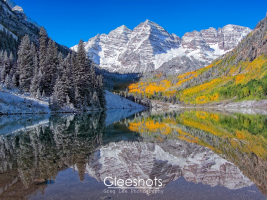 Seasons clash at the Maroon Bells in Colorado, as snowy mountain peaks contrast with vibrant aspen fall foliage in shades of orange and yellow.  Snow dusted evergreen trees also line the perfectly clear lake, which reflects the mountain scenery under a bright blue sky.   Colorado Fine Art Landscape Photography Print for Sale.