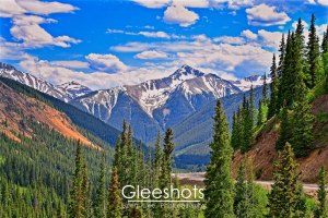 The Million Dollar Highway winds through the snow-capped San Juan mountains beneath a blue sky with scattered clouds, surrounded by dense evergreen forests.  Colorado Fine Art Landscape Photography for Sale.