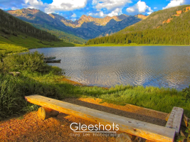 The serene Piney Lake in Colorado with a wooden bench on the shore, surrounded by lush greenery and mountains. Two rowboats are docked at the water's edge. The puffy clouds in the sky create shodows on the ground, while the setting sun casts a warm light over the landscape.  Colorado Fine Art Landscape Photography Print for Sale.