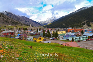 Silverton is a picturesque historic mining town in the mountains of Colorado.  Shown here are its colorful buildings framed by snowy peaks and mountains in the background. Yellow flowers dot the grassy foreground in this spring landscape.   Colorado Fine Art Landscape Photography Print for Sale.