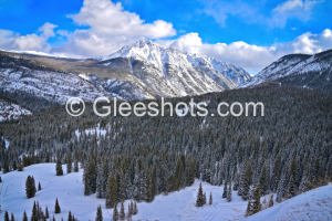 This scenic mountain winter landscape features Twilight Peak in Colorado covered in snow under a blue sky with scattered clouds. The foreground is filled with a dense, snow-dusted evergreen forest along the Million Dollar Highway. Colorado Fine Art Landscape Photography Print for Sale.