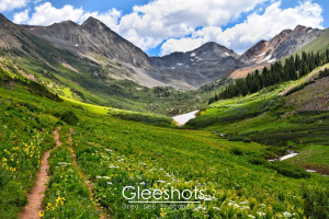 A scenic view of a lush mountain valley along Rustlers Gulch Trail, as it runs through a field of yellow and white wildflowers. The valley is surrounded by rugged mountains with patches of snow under a partly cloudy sky.  Shadows from the clouds create interesting lighting effects on the valley below.  Crested Butte Colorado Fine Art Landscape Photography Print for Sale.