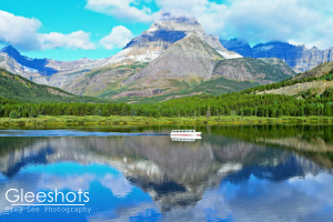 Swiftcurrent Lake reflects a crisp image of a white tour boat and rugged, snowy mountains under a blue sky scattered with clouds in Glacier National Park.  Montana Fine Art Landscape Photography Print for Sale.