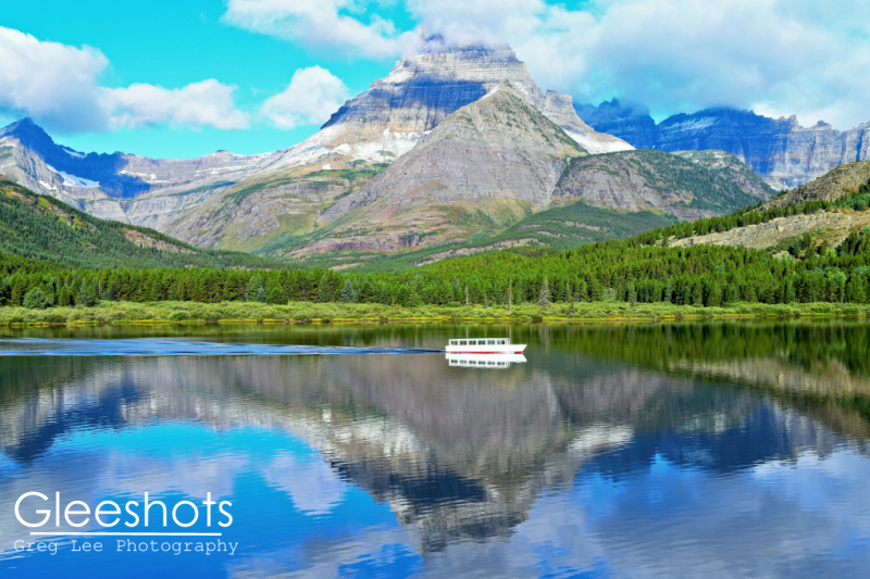 Swiftcurrent Lake reflects a crisp image of a white tour boat and rugged, snowy mountains under a blue sky scattered with clouds in Glacier National Park.  Montana Fine Art Landscape Photography Print for Sale.