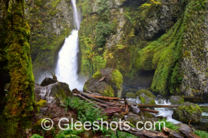 Wahclella Falls cascades down a moss-covered cliff into a rocky basin, surrounded by lush greenery and fallen logs in the Columbia River Gorge.   Oregon Waterfall Fine Art Landscape Photography Print for Sale.