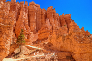 Along the Queens Garden Trail in Bryce Canyon, tall orange rock hoodoos rise against a bright blue sky.  A few people walk along the path, dwarfed by the dramatic rock formations and scattered pine trees.  National Park Fine Art Landscape Photography Print for Sale.