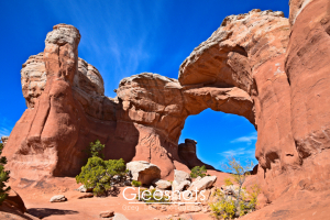 Broken Arch rises between red rock formations under a clear blue sky, with small green shrubs growing at its base at Arches National Park.  Utah Fine Art Landscape Photography Print for Sale.