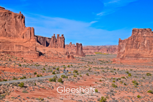 In this view of the Courthouse Towers, red rock formations and mesas rise above the desert landscape under a bright blue sky in Arches National Park. Sparse green shrubs dot the dry, reddish terrain, with a road winding through the scene.  Utah Fine Art Landscape Photography Print for Sale.