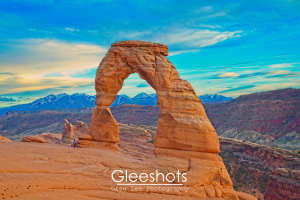 An up-close shot of Delicate Arch at sunset, with a lone person walking below.  The reds of the rock formations behind the arch can be see, with the snow-capped La Sal mountains in the distance.  Arches National Park Landscape Photography Print for Sale.