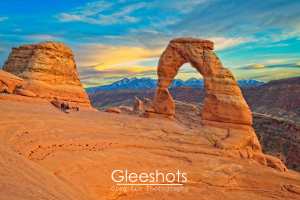 Delicate Arch in Arches National Park stands tall against a sunset sky with distant snow-capped mountains. Several people are gathered near the base, admiring the natural sandstone formation.  National Park Fine Art Landscape Photography for Sale.