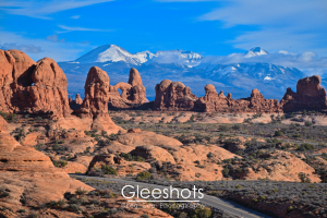 At the Garden of Eden in Arches National Park, red-orange sandstone rock formations can be seen.  The snow-capped La Sal mountains are in the distance, contrasting with a bright blue sky with low clouds.  National Park Fine Art Landscape Photography Print for Sale.