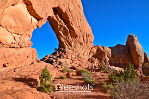 In Arches National Park, the North Window arch rises above a rocky landscape under a clear blue sky, with two people standing beneath the arch. Green shrubs dot the red terrain.   Utah Fine Art Landscape Photography for Sale.