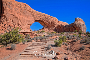 A stone stair path leads up to a large, natural red rock arch known as the North Window in Arches National Park.   A lone person can be seen walking in the distance.  Utah Fine Art Landscape Photography Print for Sale.