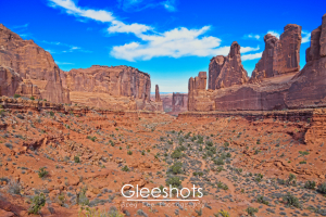 At the view of Park Avenue in Arches National Park, red rock formations and tall cliffs under a bright blue sky with wispy clouds are on display.  Sparse desert vegetation covers the rocky landscape.  Utah Fine Art Landscape Photography Print for Sale.
