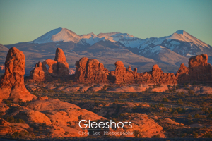 At the Garden of Eden in Arches National Park, red rock formations glow at sunset in the foreground, with snow-capped mountains in the background.  Utah Fine Art Landscape Photography for Sale.