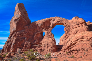Turret Arch rises against a bright blue sky at Arches National Park. Two tiny people stand beneath the arch, highlighting the massive scale of the rock formation. Shrubs dot the red, rocky landscape.  National Park Landscape Photography for Sale.