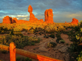 The red rock formation of Balanced Rock glows in golden sunlight at sunset under a dramatic sky at Arches National Park, Utah. A wooden fence and desert vegetation are in the foreground.  Fine Art Landscape Photography Print for Sale.