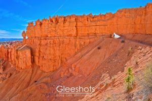 At Bryce Canyon, two people stand near a natural arch in a large orange rock formation under a blue sky. The steep, rocky slope is dotted with sparse vegetation along the Peekaboo Loop Trail.  Bryce Canyon National Park Landscape Photography Print.