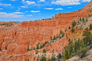 Along the Peekaboo Loop Trail leading from Bryce Point, red rock formations and cliffs rise above a forested canyon under a blue sky with scattered clouds. The rugged landscape of Bryce Canyon features distinct orange hues and sparse green pine trees.  National Park Fine Art Landscape Photography Print for Sale.
