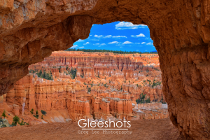 A natural rock arch frames a scenic view of orange hoodoos and cliffs under a bright blue sky in Bryce Canyon National Park.  Fine Art Landscape Photography Print for Sale.