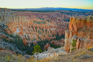 A view of Bryce Canyon taken from Bryce Point at sunset, showing dramatic orange and red rock formations called hoodoos, with a few trees scattered among the rocks and distant mountains on the horizon.  Utah Landscape Photography Print for Sale.