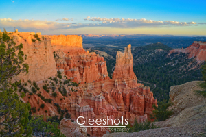 A view of Bryce Canyon at sunset from the Paria View overlook, with striking red and orange rock formations and hoodoos illuminated by golden light, set against a blue sky and distant mountains.   Utah Fine Art Landscape Photography Print for Sale.