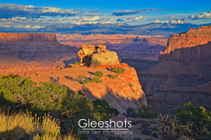 The vibrant colors of the red rocks are highlighted in this stunning sunset view of Canyonlands National Park.   Layered red rock formations, green shrubs, and distant snow-capped mountains under a blue sky are shown, with two people walking on the rocky outcropping below.   Utah Fine Art Landscape Photography Print for Sale.