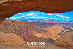 In this scene through Mesa Arch at Canyonlands National park in Moab, the red rock canyon valley below is highlighted in shadows and sunlight along with a bright blue sky scattered with clouds.   Utah Fine Art Landscape Photography Print for Sale.