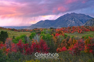 A vibrant autumn landscape in Cascade Meadows with colorful red, orange, and green trees, rolling hills, and Mt Timpanogos in the background.  A dramatic purple sunset sky with clouds and rain can be seen, as rain falls over Utah valley in the distance.   Fine Art Landscape Photography Print for Sale.