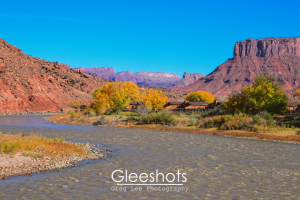 The Colorado River flows through a rocky desert landscape in Moab, with autumn trees and red cliffs under a clear blue sky; small cabins nestle among the trees.  Utah Fine Art Landscape Photography for Sale.