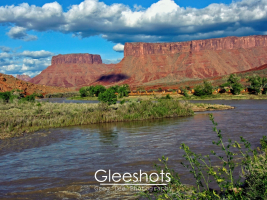 The Colorado River flows in the foreground with green vegetation along its banks; behind it are red rock mesas and cliffs under a partly cloudy blue sky in Moab.  Utah Fine Art Landscape Photography for Sale.