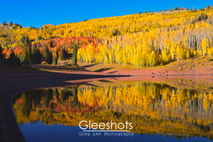 Desolation Lake reflects a hillside covered in vibrant autumn trees with yellow, orange, and green foliage under a clear blue sky in Big Cottonwood Canyon.  Utah Landscape Photography Print for Sale.