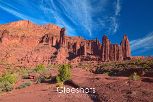 The red and orange rock formations of the Fisher Towers rise dramatically against a bright blue sky with wispy clouds, surrounded by desert vegetation and rugged terrain near Moab.  Utah Fine Art Landscape Photography for Sale.