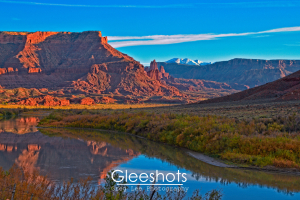 An autumn sunset view of red rock cliffs and the Fisher Towers in Moab, with snow-capped mountains in the background.  The mesas reflect in the Colorado River, bordered by green and yellow foliage.  Utah Fine Art Landscape Photography Print for Sale.