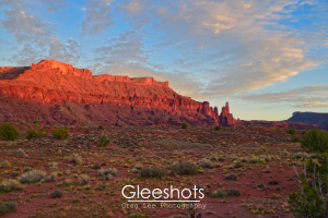 The red rocks of the mesas around Fisher Towers glow red at sunset,  with scattered clouds overhead and desert shrubs in the foreground.  Utah Fine Art Landscape Photography Print for Sale.