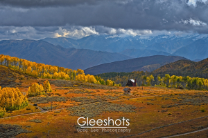 On Guardsman Pass, filtered sunlight shines on two small buildings and golden autumn trees among rolling hills, with dramatic mountain peaks and dark clouds in the background.   Utah Fine Art Landscape Photography for Sale.